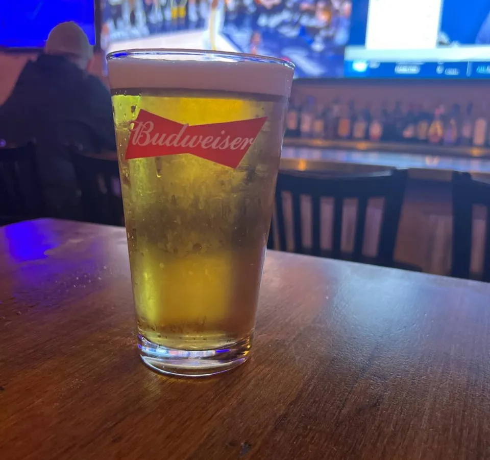 A cold Budweiser beer in a pint glass sits on a wooden bar table with a sports game displayed on a blue-lit screen in the background.