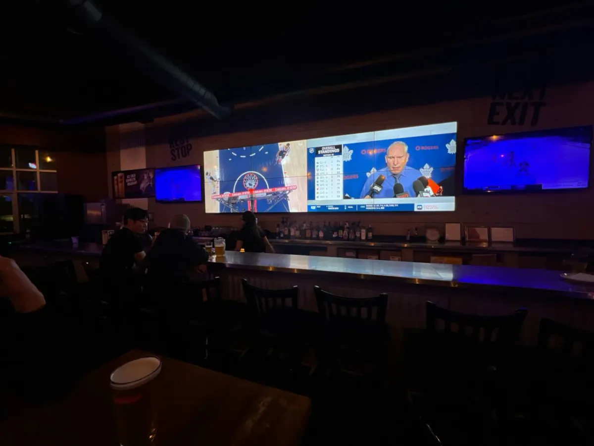 A sports bar interior with multiple large screens displaying sports content and a man speaking at a podium during what appears to be a press conference or broadcast.